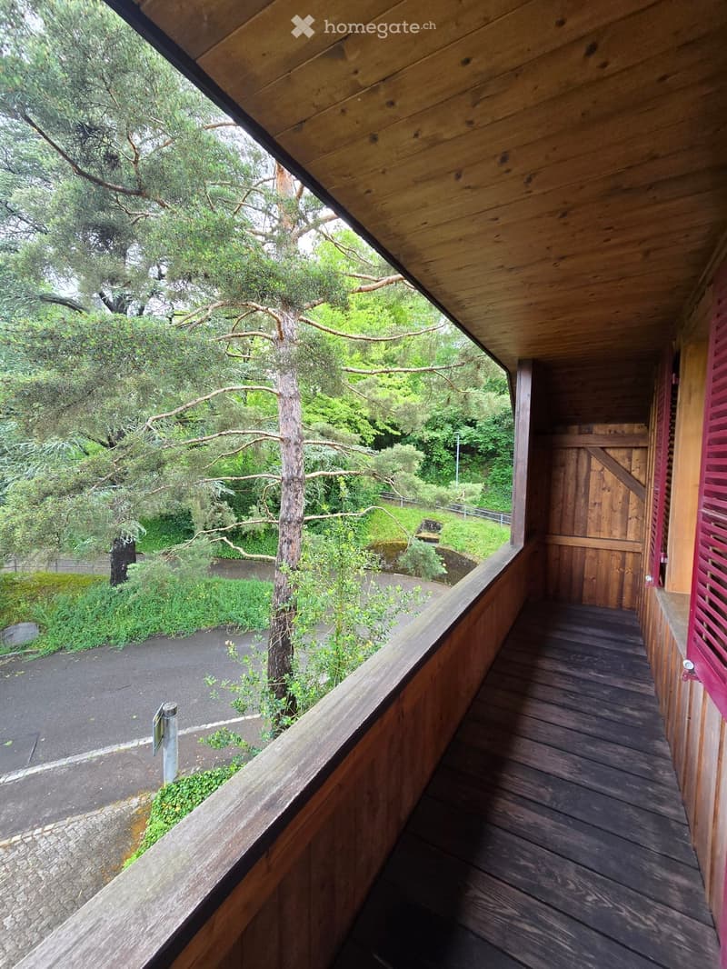 Wooden balcony with railing, leading to a wooden door, surrounded by greenery and trees.