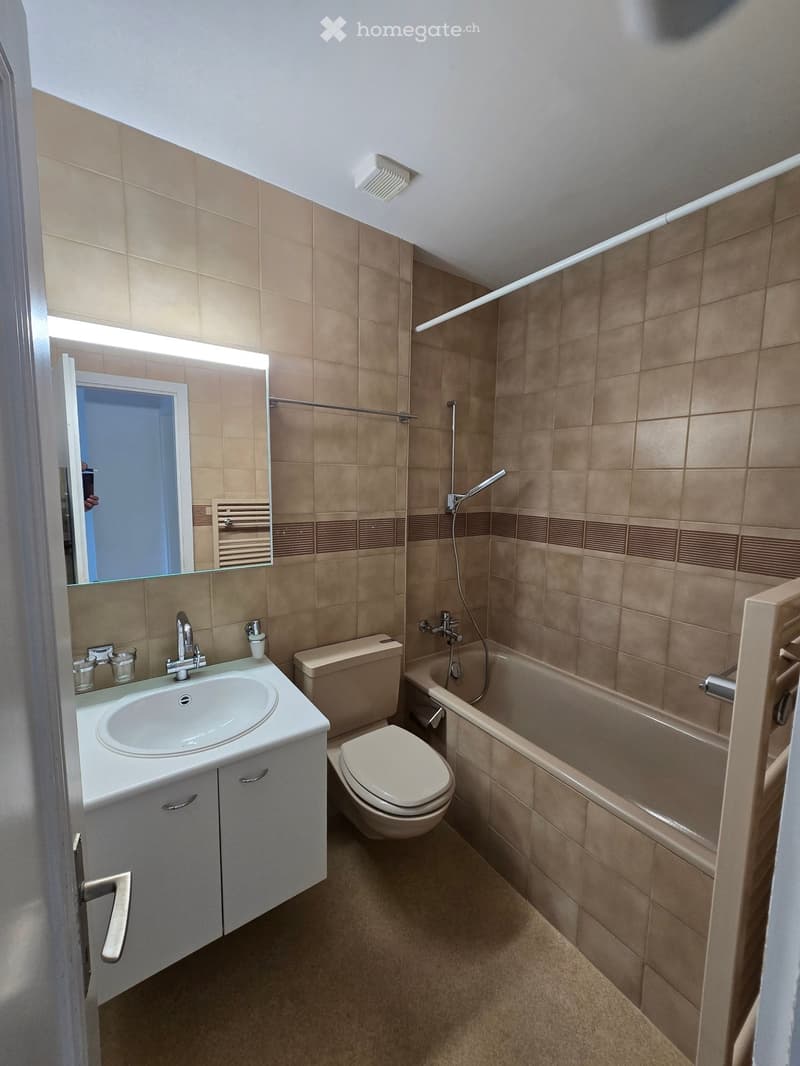 Modern bathroom with beige tiles, a white sink with a mirror, a toilet, a bathtub, and a towel rack.