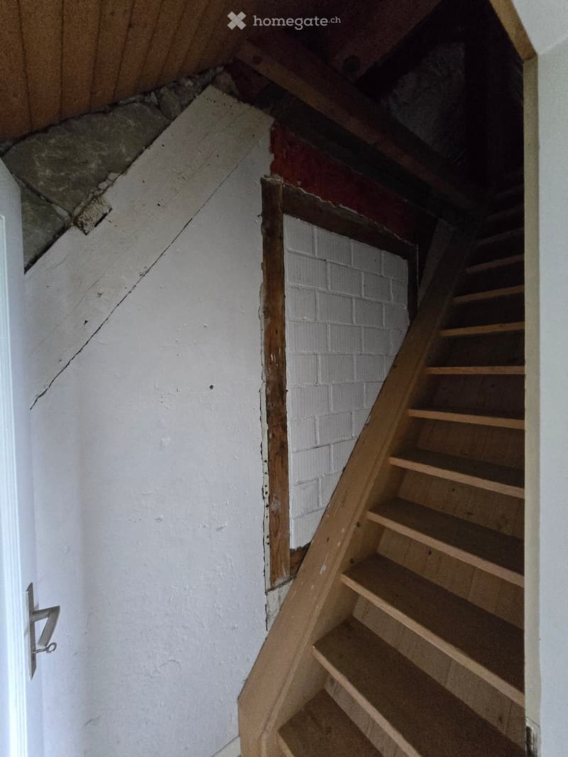 Wooden staircase leading up, white walls, exposed brick section, unfinished ceiling with visible beams and insulation.