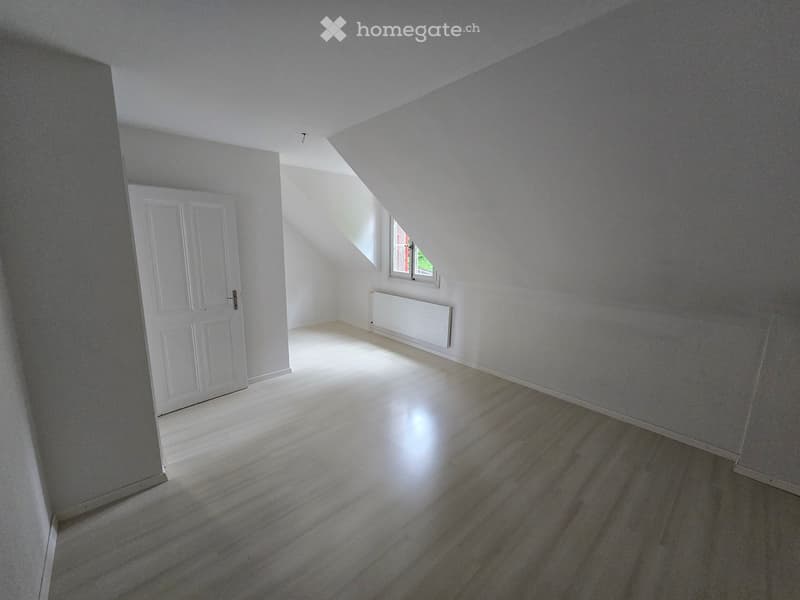 Empty attic room with wooden flooring, white walls, a white door, and a small window letting in natural light.