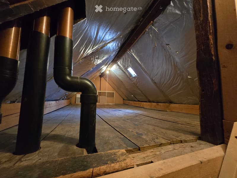 An attic space with wooden flooring, exposed wooden beams, insulation, and a black pipe running through it. The area is unfinished and appears to be under construction or renovation.