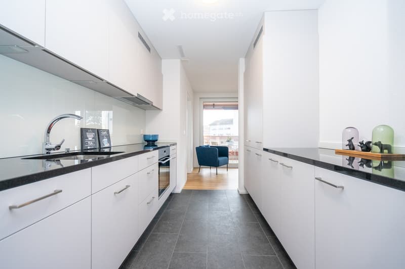 Modern kitchen with white cabinets, black countertops, stainless steel sink, and a view into a living area with a blue armchair and small table.