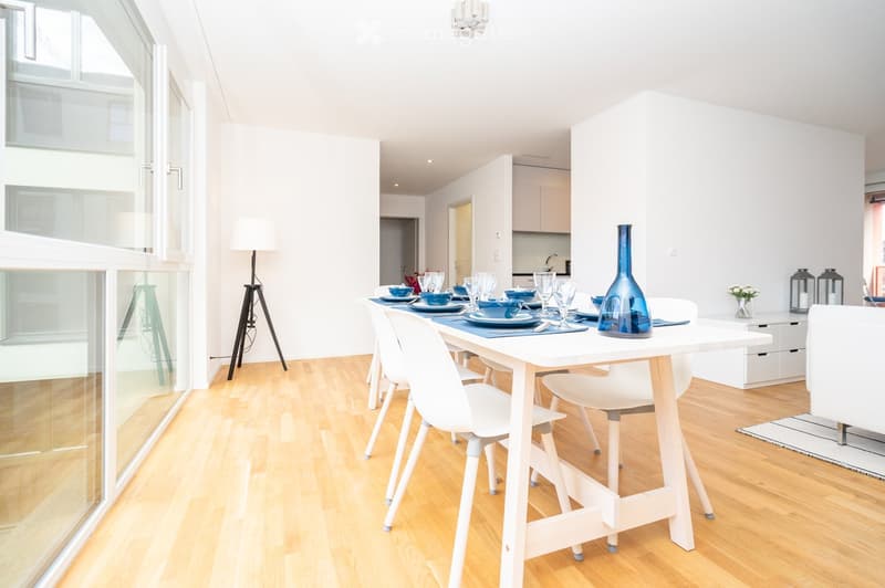 Bright dining area with a white dining table, white chairs, and a blue vase on the table. The room features wooden flooring, large windows, and a modern lamp in the corner.