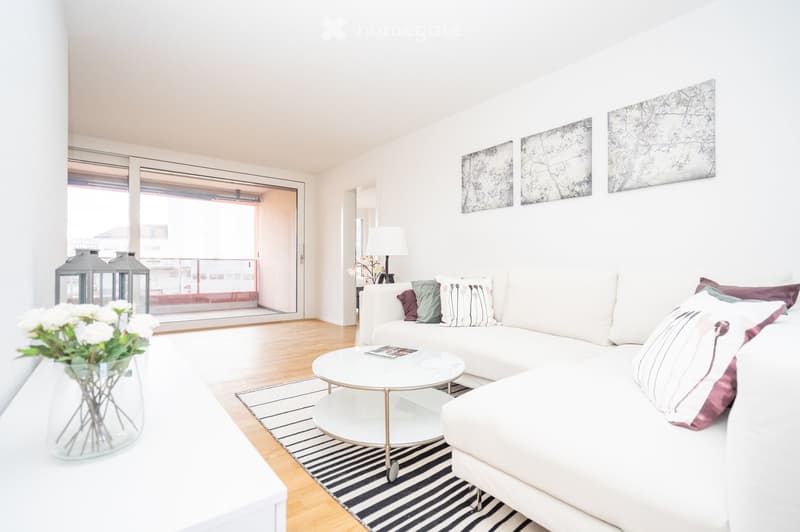Modern living room with white walls, light wooden flooring, a white sectional sofa with colorful pillows, a white coffee table, a floor lamp, and a vase with white flowers on a white console table. There is a large window with a view of a balcony.