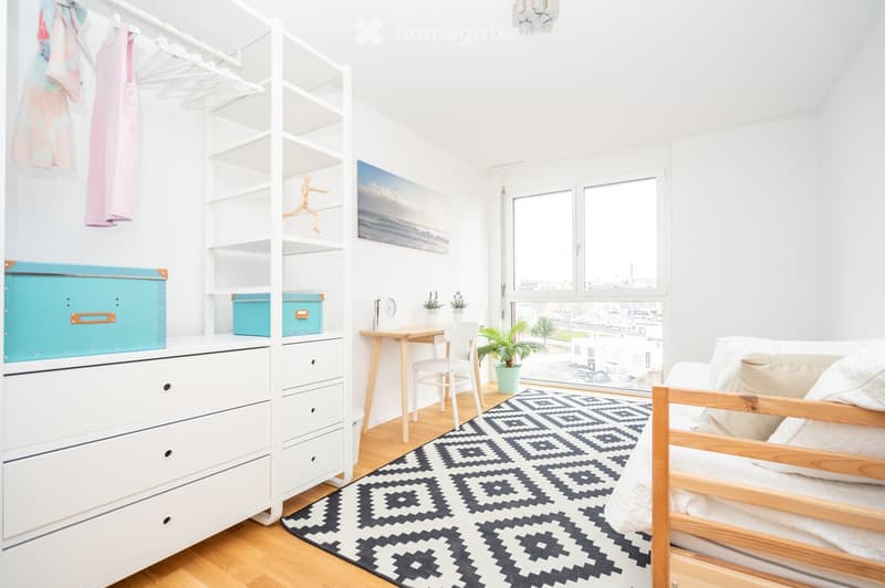 Bright bedroom with a wooden bed, white chest of drawers, built-in shelving unit, desk with chair, and a patterned rug.