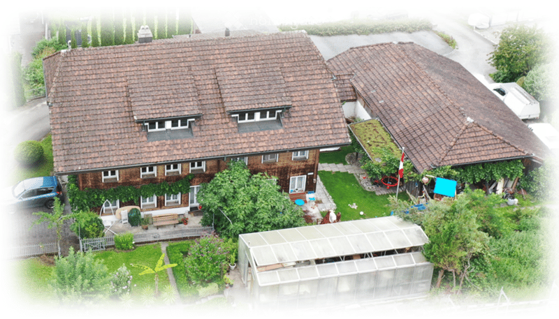 Aerial view of a two-story house with a tiled roof, surrounded by a well-maintained garden, a greenhouse, and a parked car.