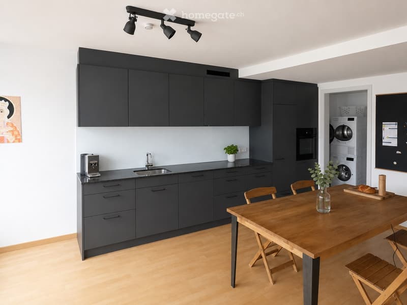 Modern kitchen with dark grey cabinetry, a central island, a sink, a coffee maker, and a wooden dining table with chairs. A washing machine and dryer are visible in an adjacent area.