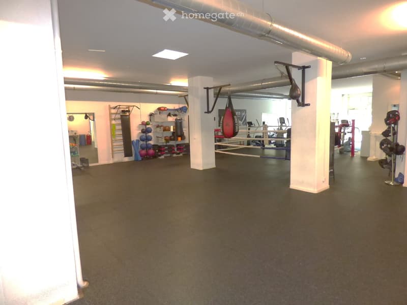Well-lit gym space with boxing ring, punching bag, weight shelves, mirrors, and exercise bikes.