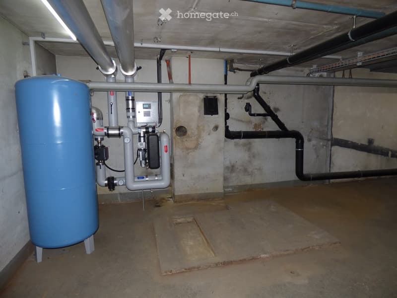 Utility room with exposed pipes, a water tank, and control panel. Concrete walls and floor.
