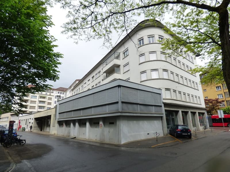 Modern multi-story building with white facade, large windows, and balconies. Features a flat, extended lower section with garage entrances and parking spaces.