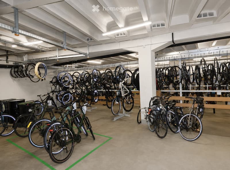 A spacious indoor area filled with numerous bicycles hanging from the ceiling and standing on the floor, with a clean, industrial-style design and exposed metal beams.