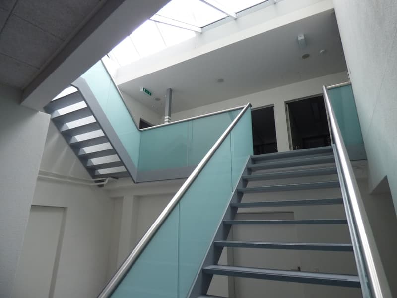 Modern interior staircase with grey steps, glass railings, and a skylight above, leading to an open doorway.