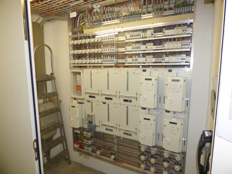 Electrical panel with circuit breakers, meters, and pipes with valves, mounted on a wall with a metal ladder nearby.