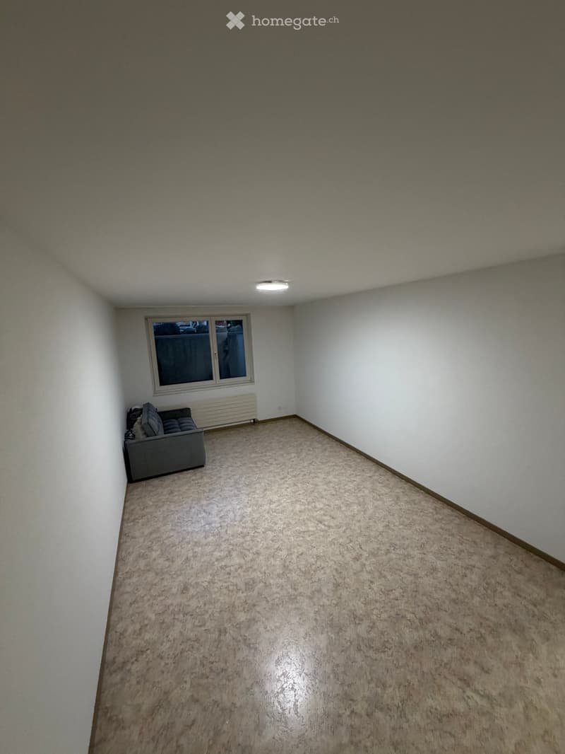 A small, unfurnished room with a single sofa, a window, and a radiator. The walls are white, and the floor is carpeted.