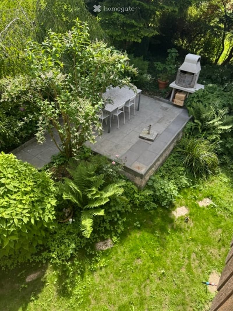 A small, well-maintained garden with a raised patio area featuring a dining table and chairs, surrounded by lush greenery and trees.