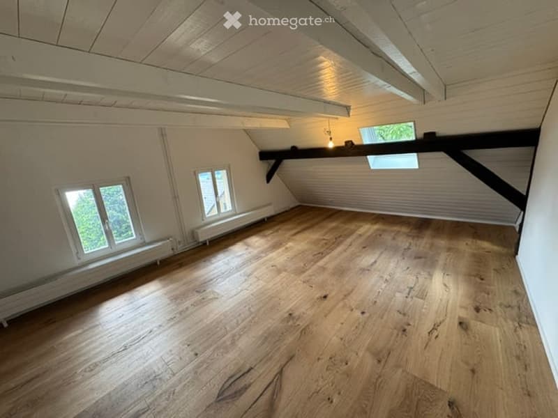 Empty attic room with wooden flooring, white walls, exposed wooden beams, and two windows letting in natural light.