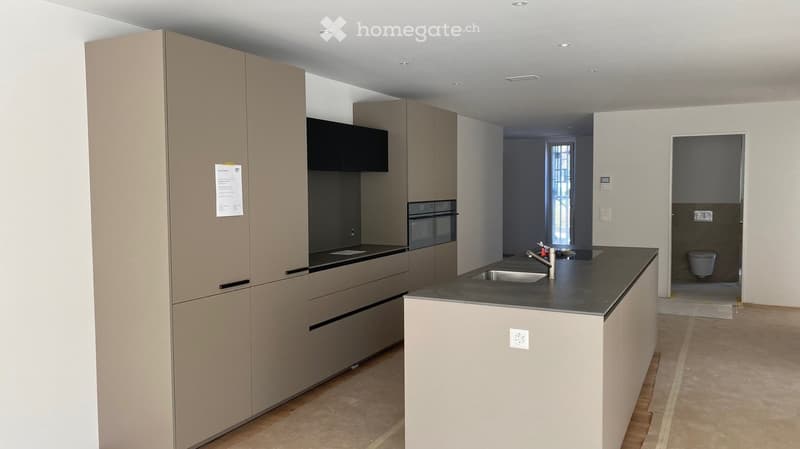 Modern kitchen with light-colored cabinets, a black countertop, and a sink. An oven is visible, and a small bathroom is partially visible through an open doorway.