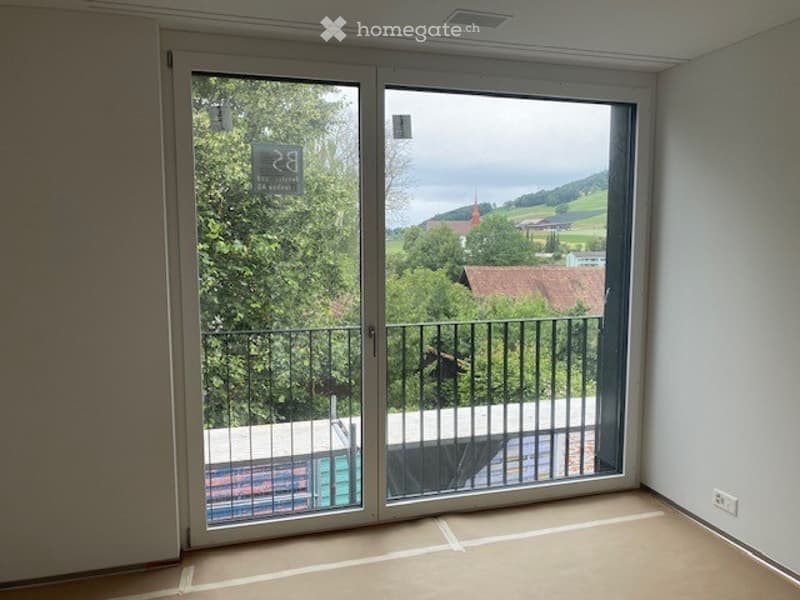 Empty room with large sliding glass doors opening to a balcony, showing a scenic view of greenery and a town with a church tower.