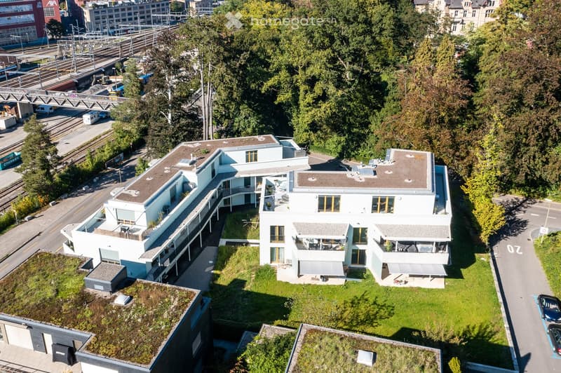 Aerial view of a modern, multi-story residential building with white walls, large windows, and green rooftop areas. The building is surrounded by trees and has a landscaped garden area in front.
