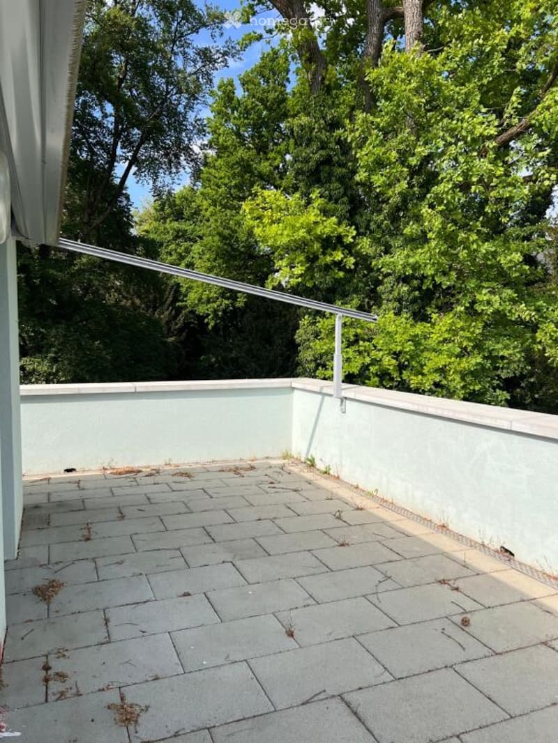 Empty balcony with light blue walls, tiled floor, metal railing, and lush green trees in the background.