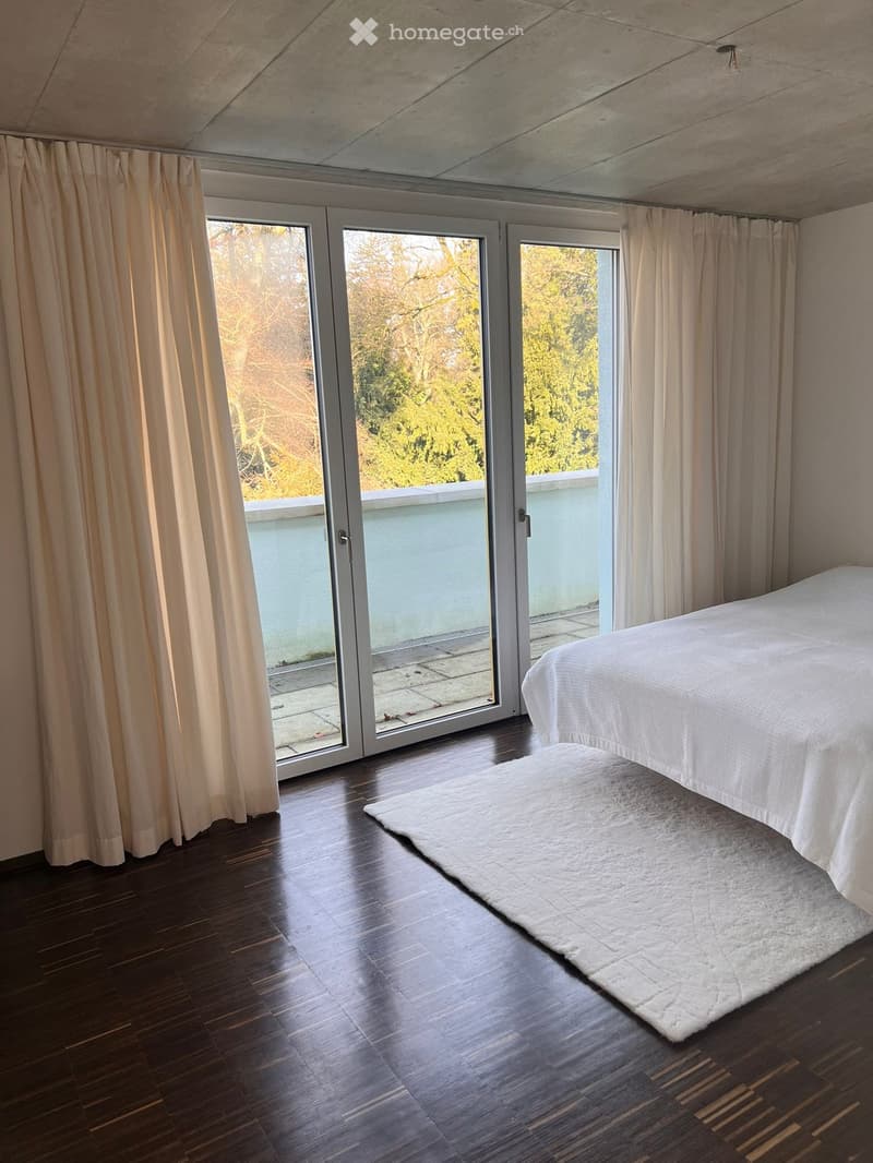 Modern bedroom with wooden flooring, large sliding glass doors leading to a balcony, and a white bed with a shaggy rug.