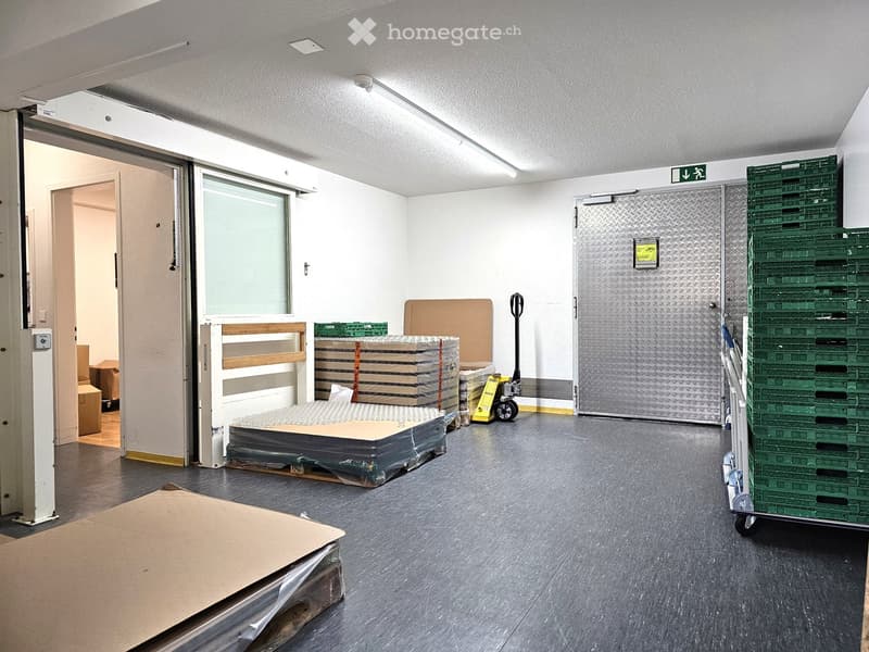 Empty storage room with grey flooring, white walls, and large industrial doors. Stacks of cardboard boxes and green crates are visible, along with a yellow pallet jack.