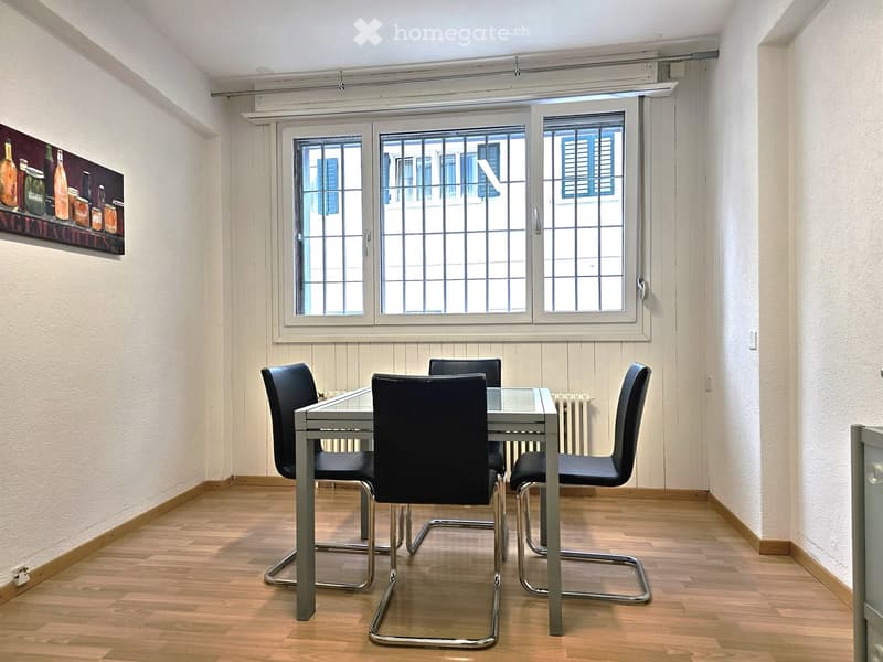 Dining room with a glass table and four black chairs, wooden flooring, and large windows with shutters.