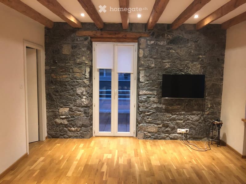 Empty room with exposed wooden beams, stone walls, wooden flooring, and a large window with blinds.
