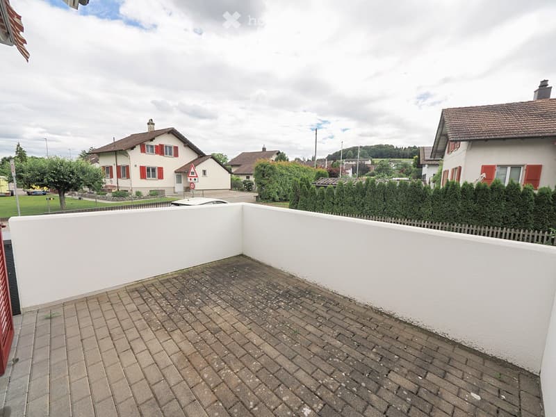 Brick-paved balcony with white walls, overlooking a residential area with houses, trees, and a parked car.