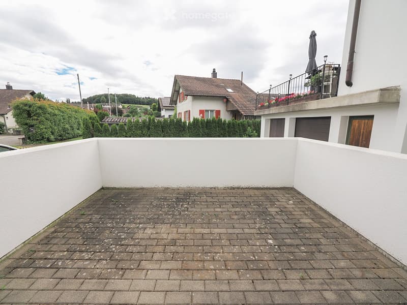 Empty brick-paved balcony with white walls, overlooking a residential area with houses and greenery.
