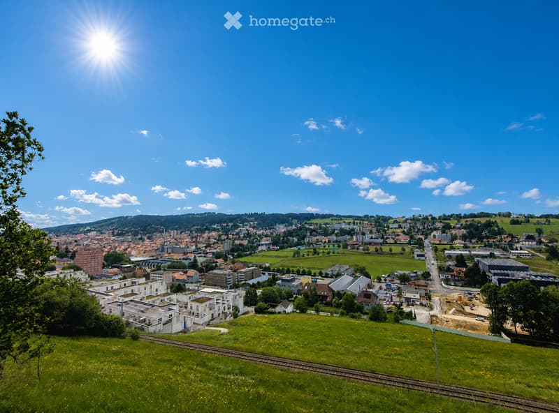 A panoramic view of a town with a mix of residential and commercial buildings, surrounded by green fields and hills under a clear blue sky.