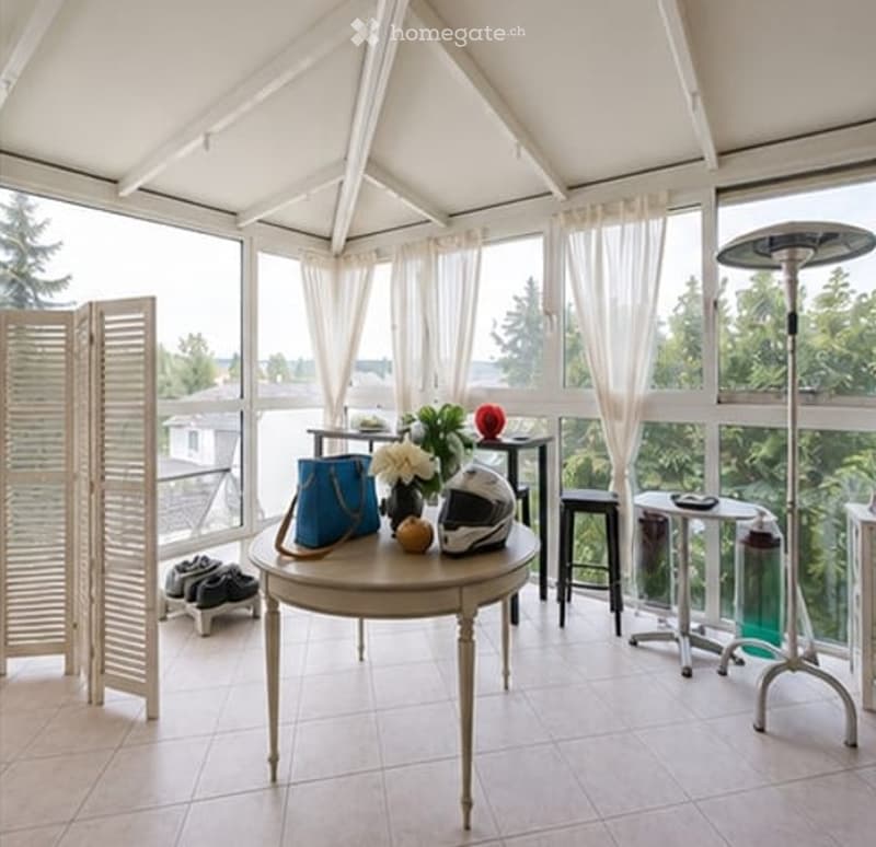 Bright, airy room with large windows and a view, furnished with a round table, stools, a chair, and decorative items.