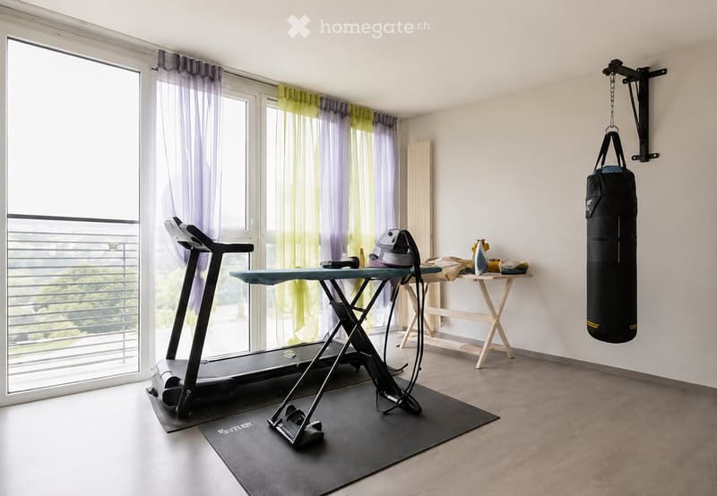 Well-lit room with large windows and curtains, featuring a treadmill, ironing board, small table with folded clothes, and a punching bag hanging on the wall.
