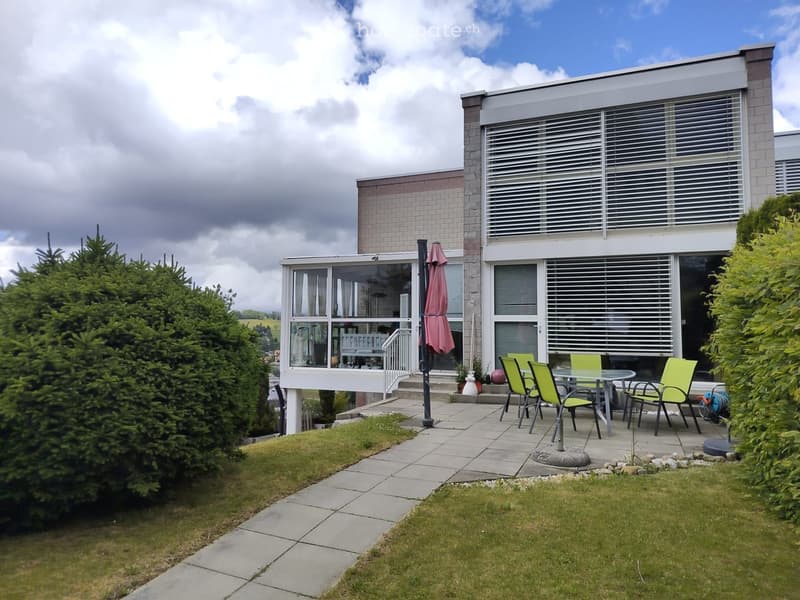 Outdoor patio area with a glass-walled extension of the house, featuring a table and chairs, surrounded by greenery and a paved walkway.