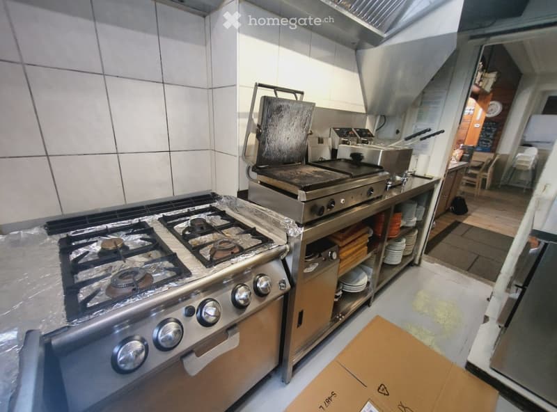 Commercial kitchen with stainless steel appliances including a gas stove, griddle, and oven. Shelves hold plates and utensils, with a cardboard box on the floor.