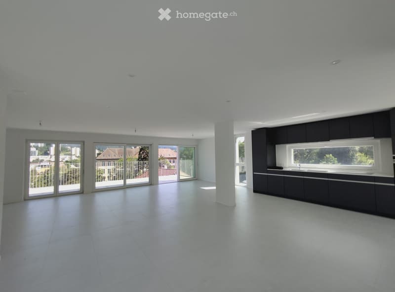 Open-plan unfurnished space with white tiled flooring, large sliding glass doors leading to a balcony, and a modern kitchen area with black cabinetry and a white countertop.