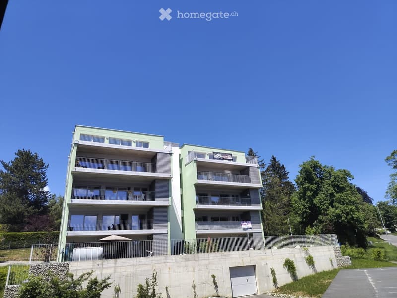 Modern multi-story apartment building with balconies, green facade, and a raised ground floor with a garage entrance.