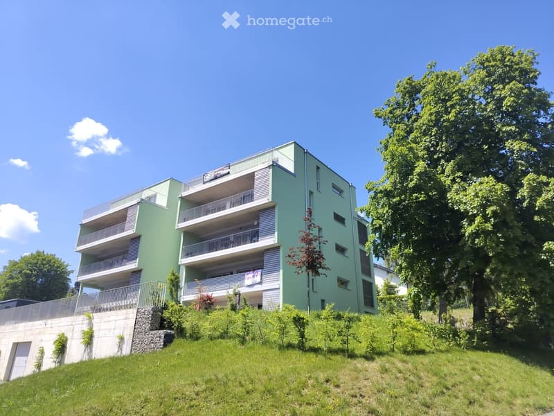 Modern multi-story apartment building with green and white exterior, multiple balconies, surrounded by trees and greenery.