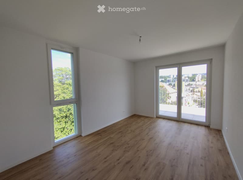 Empty room with wooden flooring, white walls, large windows, and sliding glass doors leading to a balcony.