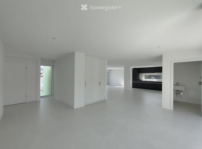 A completely empty room with white walls, a white tiled floor, and a large open space. The room features a white closet, a doorway leading to another room, and a small kitchen area visible in the background.