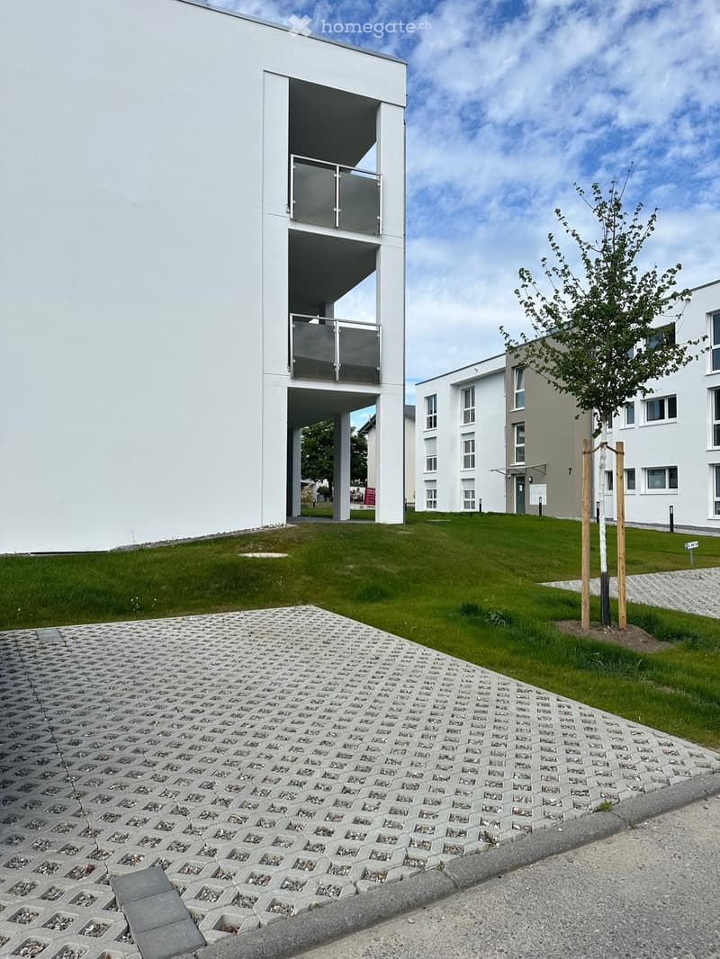 Modern white building with a raised ground floor, glass balcony, and surrounding green lawn.