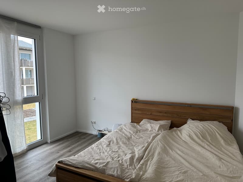 A modern bedroom with a wooden bed, headboard, and nightstand. The room features light-colored walls, wooden flooring, and a large window with a view of the outside.