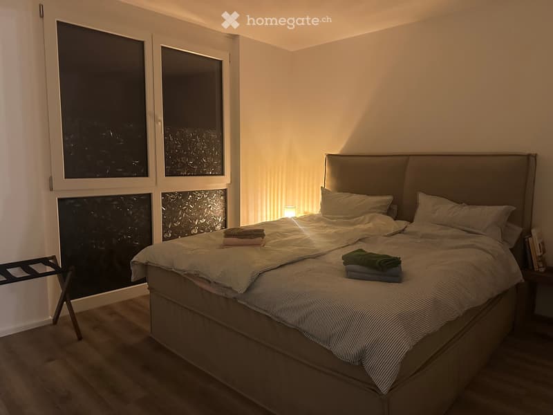 Modern bedroom with a large bed, nightstand, and a small chair near the window. The room features large windows with a view and warm lighting.