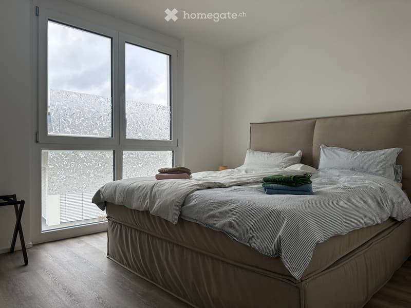 Modern bedroom with large windows, a neatly made bed with pillows, a headboard, and folded towels on the bed.