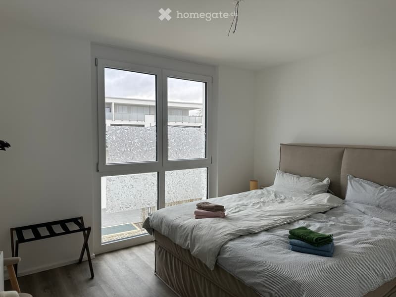 Modern bedroom with a large bed, neatly made with pillows and blankets, a nightstand with a lamp, and a radiator cover near the window. The room features large windows with a view of a balcony and adjacent building.