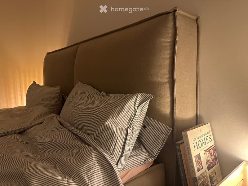 A neatly made bed with a headboard, pillows, and a striped blanket, accompanied by a stack of books on a bedside table.