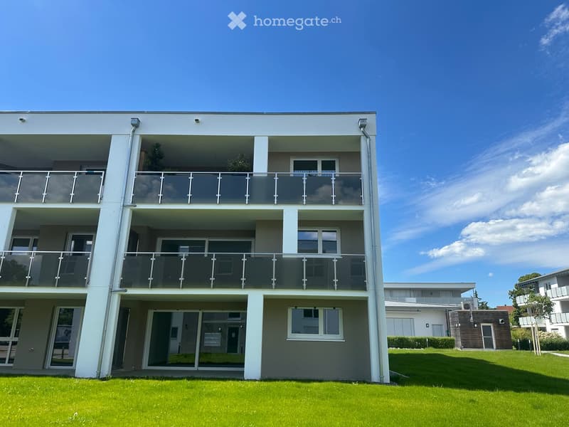 Modern two-story building with large glass balconies and white exterior walls, surrounded by green lawn.