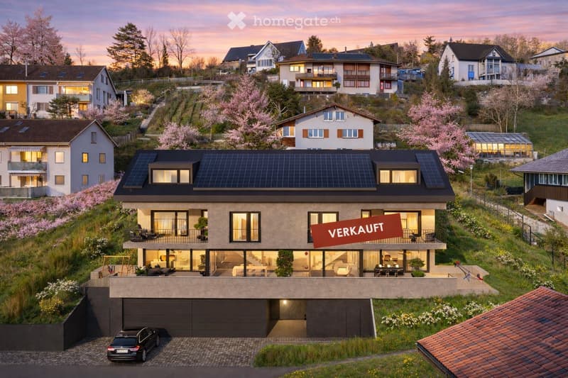 Modern two-story house with solar panels on the roof, large windows, and a spacious balcony. The house is surrounded by a landscaped garden with blooming trees and shrubs. A car is parked in the driveway, and the house is marked as "VERKAUFT" (sold).