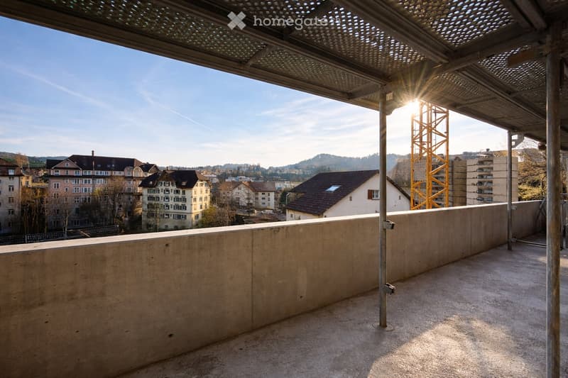 Empty balcony with concrete floor and low wall, metal railing, and a perforated metal roof providing partial shade. The view shows a cityscape with residential buildings and a construction crane in the distance.