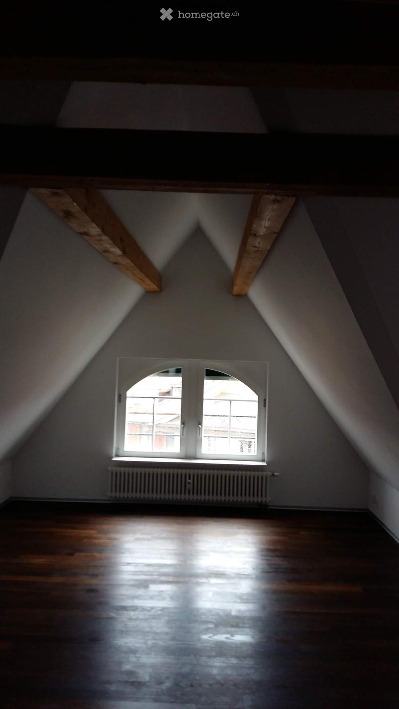 Unfurnished Room. Empty attic room with wooden floors, slanted ceilings, and a single window letting in natural light. The room features exposed wooden beams and a radiator below the window.
